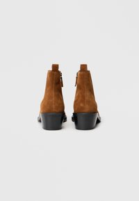 Braune Wildleder-Stiefeletten mit einem niedrigen schwarzen Absatz, ausgestattet mit einem seitlichen Reißverschluss und einer Lasche zum einfachen Anziehen. Glatte Textur und schlichtes Design.