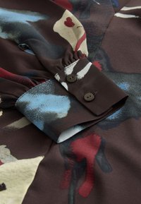 Blouse met lange mouwen in donkerbruine stof met abstracte patronen in blauw, rood en crème. Voorzien van een knoopmanchet en details met plooien aan de mouwen.