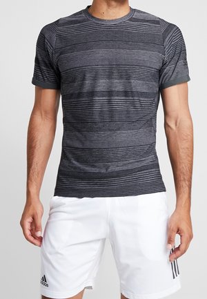 Męski tułów ubrany w dopasowaną, ciemnoszarą koszulkę w paski z krótkim rękawem oraz białe sportowe spodenki Adidas na jednolitym tle.