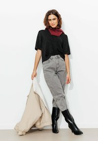 Haut en maille noir à manches courtes, jean gris avec un motif subtil, bottes noires montant jusqu'au genou, tenant une veste beige doublée de blanc.