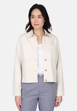 Femme aux cheveux foncés portant une veste crème à boutons, une chemise blanche et un pantalon rayé bleu et blanc, debout devant un arrière-plan uni.