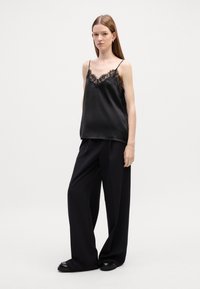 Svart sateng-camisole med blonderkant, kombinert med vide svarte bukser og kraftige svarte sko. Modellen står mot en ensfarget bakgrunn.