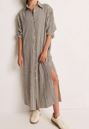 Woman wearing a long beige dress with black vertical stripes, buttons down the front, side slit, rolled-up sleeves, and white sneakers.