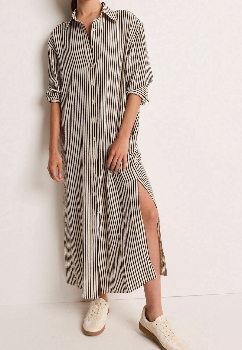 Femme portant une longue robe beige à rayures verticales noires, boutons devant, une fente sur le côté, manches retroussées et baskets blanches.
