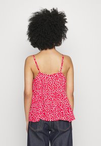 Donna con capelli ricci che indossa un top senza maniche rosso con pois bianchi e jeans blu scuro, in piedi su uno sfondo semplice.