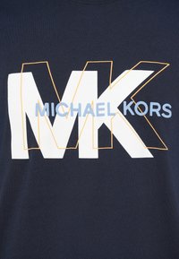 Tejido azul marino con grandes letras blancas "MK" contorneadas en naranja, superpuestas con "MICHAEL KORS" en texto en mayúsculas de color azul claro.