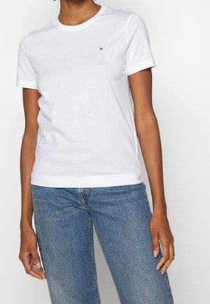 Persoon in een eenvoudige witte T-shirt met korte mouwen met een klein logo op de borst en blauwe spijkerbroek, staande voor een witte achtergrond.