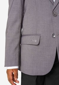 Zbliżenie na osobę w szarym żakiecie z klapkową kieszenią i widocznymi guzikami, noszącą białą koszulę i czarne spodnie.