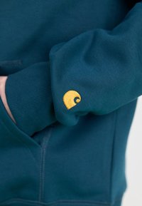 Felpa verde petrolio con polsini a coste. Presenta un piccolo logo giallo ricamato sul polsino sinistro. Il tessuto ha una finitura morbida e testurizzata.