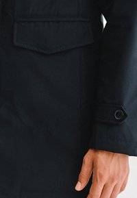 Granatowa kurtka o gładkiej fakturze, z kieszenią na piersi, zapięciem na guziki przy mankietach oraz widocznymi detalami szwów.