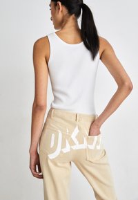 Top a maniche lunghe bianco a coste abbinato a pantaloni beige con grandi testi grafici bianchi sui taschini posteriori. Modello mostrato da dietro.