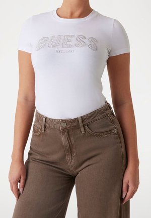 White cotton T-shirt with short sleeves featuring a sparkling "GUESS EST. 1981" logo, paired with high-waisted brown denim jeans.