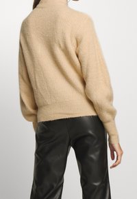 Beige gestrickter Pullover mit hohem Kragen, gerippter Textur, Ballonärmeln und schmalem Saum, kombiniert mit schwarzen Lederhosen.