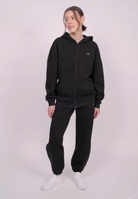Un sweat à capuche noir avec une fermeture éclair, des poches à l'avant et un petit logo ; associé à un pantalon de jogging noir assorti et des baskets blanches. Texture de tissu lisse.