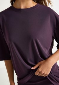 Fialové oversized tričko s krátkými rukávy, kulatým výstřihem a hladkou texturou. Ruka drží látku na boku. Jednoduchý design, bez vzorů.