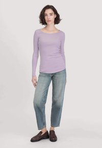 Lichtpaarse long-sleeve t-shirt gecombineerd met lichtblauwe relaxte jeans en donkergouden instappers, tegen een neutrale achtergrond.