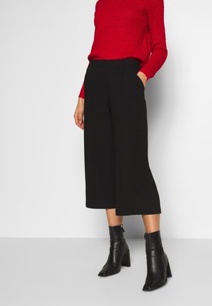 Culottes noires en tissu lisse, avec des plis sur le devant et des poches sur les côtés, associées à un pull rouge texturé et des bottines noires.