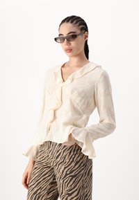 Cremefarbende Bluse mit Rüscheneinsatz und ausgestellt geschnittenen Ärmeln, kombiniert mit beigen, zebra-gestreiften Hosen. Die Bluse hat ein strukturiertes Blumenmuster.