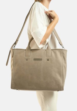 Femme portant une chemise blanche et un grand sac fourre-tout beige avec poignées et bandoulière sur l’épaule.