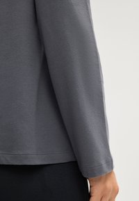 Šedá košile s dlouhým rukávem vyrobená z hladkého bavlněného směsi. Obsahuje prošité lemy a přiléhavé manžety, s jednoduchým designem bez vzorů.