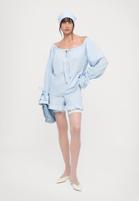 Woman wearing light blue off-shoulder blouse, matching shorts with lace trim, white headscarf, holding blue woven clutch, and white pointed shoes.