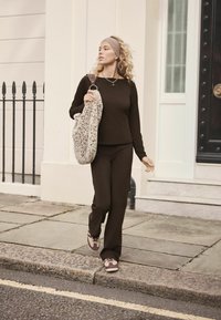 Brauner Langarm-Oberteil und passende Hose, kombiniert mit gemusterten Turnschuhen. Sie hält eine gepunktete Tasche und geht die Straße einer Stadt entlang.