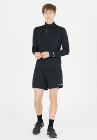 Sort langærmet atletisk top med lynlås krave og grå detaljer, parret med matchende sorte shorts med logo. Båret med sorte sneakers.