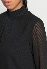 Schwarze Bluse mit hohen Kragen und strukturiertem Zickzackmuster, mit durchsichtigen langen Ärmeln und einem gerafften Kragen.