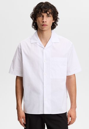 Młody mężczyzna o kręconych ciemnych włosach, ubrany w prostą białą koszulę z krótkim rękawem i czarne spodnie na neutralnym tle.