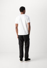T-shirt blanche en coton à manches courtes, associée à un pantalon cargo noir avec des poches, et des baskets noires avec une semelle blanche.