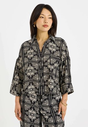 Femme aux cheveux noirs raides portant une chemise à manches longues et un pantalon assortis à motif floral noir et blanc, boucles d'oreilles créoles dorées et bracelets.