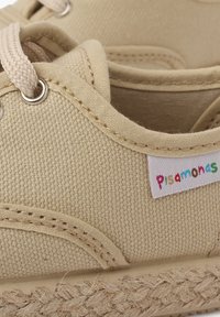 Béžové plátenné tenisky s okrúhlou špičkou, textúrovaným zvrškom a pletenou jutovou podrážkou. Obsahujú kovové očká a farebný �štítok s logom po strane.