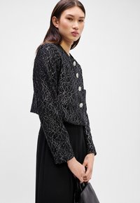Giacca a crop in pizzo nero con motivi floreali metallici, bottoni rotondi e finitura testurizzata. Abbinata a una gonna nera e una borsa.