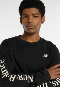 Mustapaita, jossa on pitkät hihat ja joka on valmistettu puuvillasta. Rinnassa on valkoinen logo ja vasemmassa hihassa kermanväriset tekstit "New Balance".