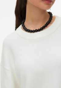 Collier noir en perles de pierres brillantes, présentant une forme ronde et uniforme. Porté sur un pull léger en tricot blanc.