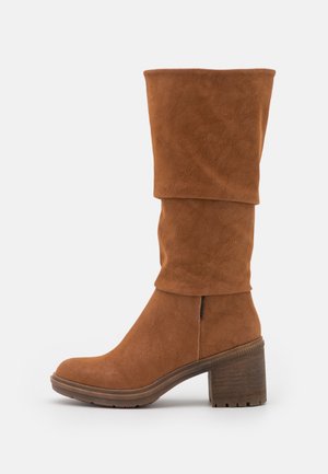 Ruskeat mokkanahkaiset puolivahteiset saappaat, joissa löysä varsi, pyöreä kärki, paksu puisella korolla varustettu paljas pohja ja kumipohja valkoisella taustalla.