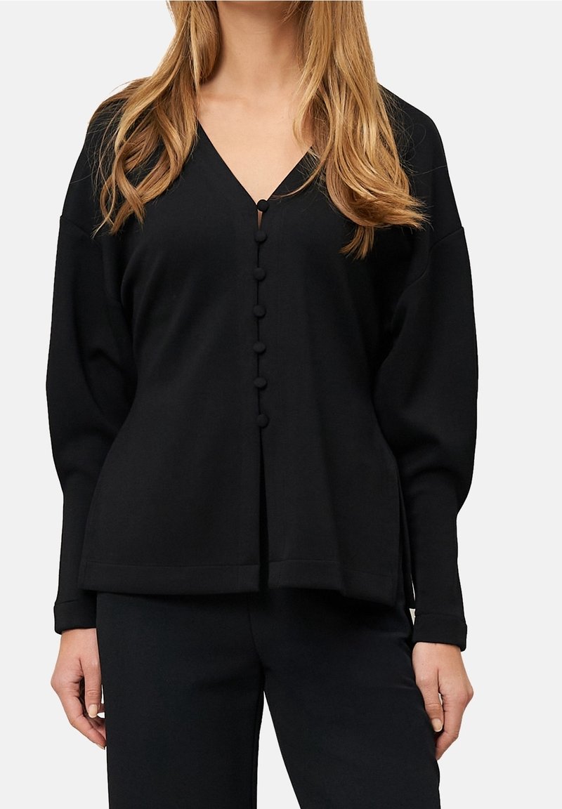 Femme portant un cardigan noir ample à col en V boutonné avec des manches longues et un pantalon noir assorti.