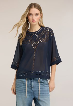 Jeune femme portant un chemisier bleu marine transparent avec des découpes géométriques et des manches trois-quarts, associé à un jean bleu clair.