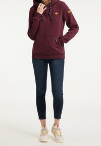 Sweat à capuche marron avec poche kangourou, capuche réglable par cordon et logo parapluie jaune. Associé à un jean skinny bleu foncé et des baskets.