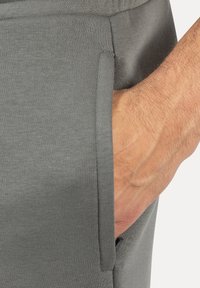 Pantalones deportivos grises en una tela suave, con la mano en el bolsillo lateral; presentan una textura suave y un detalle de costura mínimo.