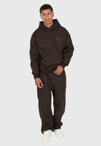 Giovane in piedi che indossa un felpa marrone scuro e pantaloni abbinati con il testo "Dropsize", scarpe da ginnastica bianche, su uno sfondo chiaro uniforme.