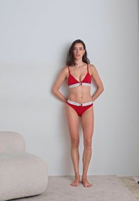 Punainen bikinitsetti, jossa on kolmion muotoinen bralette ja korkea leikkaus alushousut. Korostettu valkoisilla logonauhoilla. Sujuva kangas, minimalistinen muotoilu.