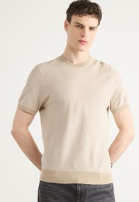 Beige rövidujjú kötött pulóver, kerek nyakkal, bordás mandzsettával és texturált felülettel. Az alja egy sima bordás szalaggal rendelkezik.