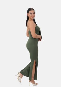 Vestido ajustado verde oliva con tirantes finos, aberturas laterales y textura acanalada. Combinado con sandalias de tacón blancas con un diseño trenzado.