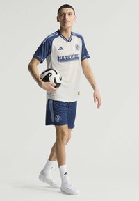 Tenue de football comprenant un maillot rayé blanc et bleu foncé avec logo, et un short assorti. Le joueur tient un ballon de football noir et blanc.