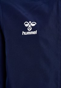 Marineblå atletisk jakke med en glat overflade, der har et hvidt Hummel-logo. Enkel design uden yderligere mønstre eller accenter.