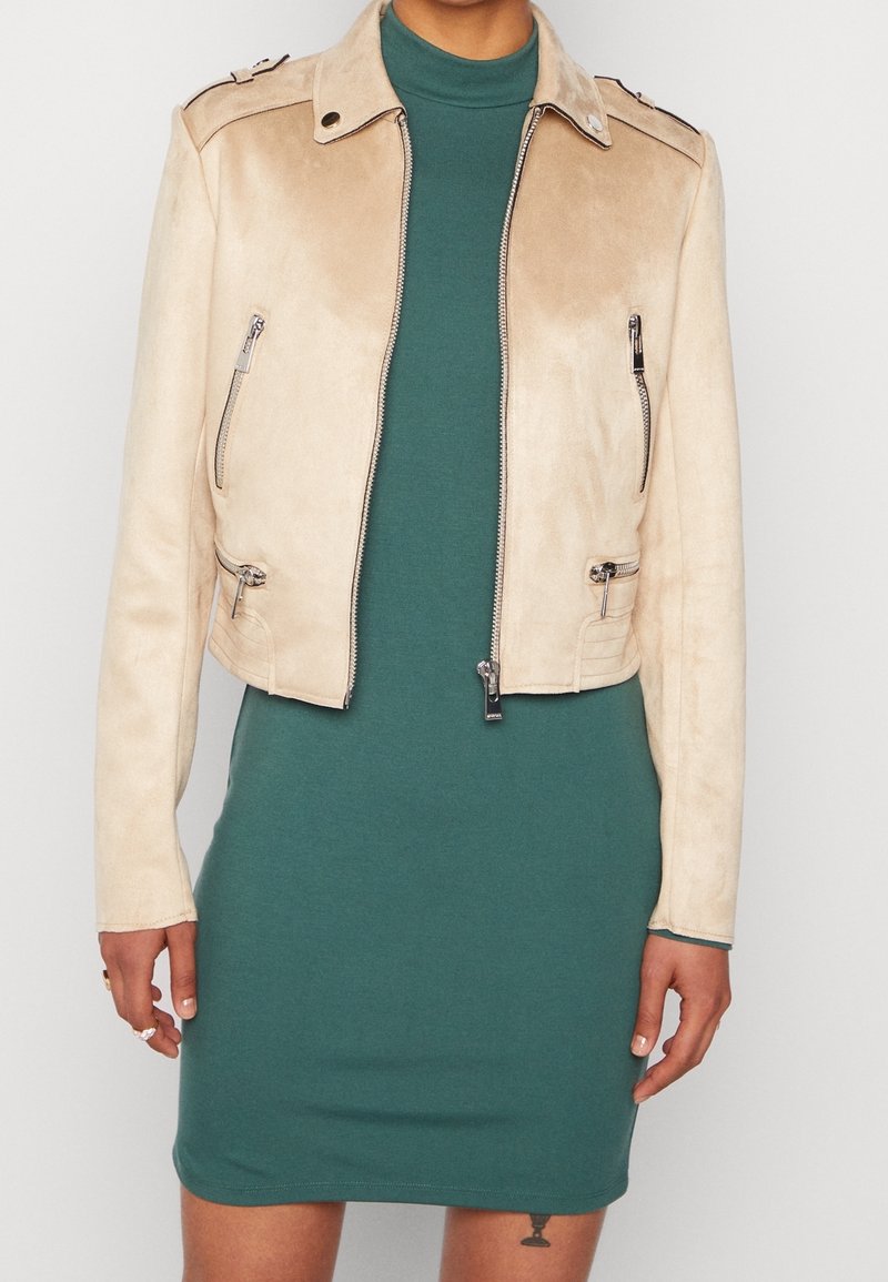 Veste cropped en suède beige avec poches zippées, épaulettes, et finitions en métal argenté, portée sur une robe ajustée teal.