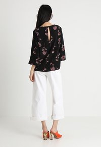 Schwarzes Hemd mit Blumenmuster, Glockenärmel und Keyhole-Rücken-Detail, kombiniert mit weißen weiten Hosen und orangefarbenen High Heels.