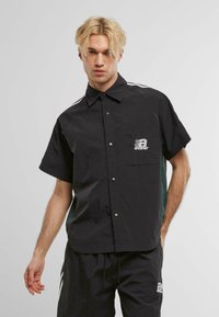 Camisa negra de manga corta con un cierre frontal de botones, que presenta un bolsillo en el pecho y un logotipo blanco. Paneles laterales en verde oscuro y rayas blancas.