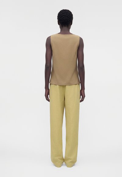 Άτομο με κοντά, σγουρά μαλλιά στέκεται με την πλάτη προς τα εμπρός, φορώντας ένα αμάνικο καφέ τοπ και άνετο ανοιχτό κίτρινο παντελόνι μπροστά σε απλό φόντο.